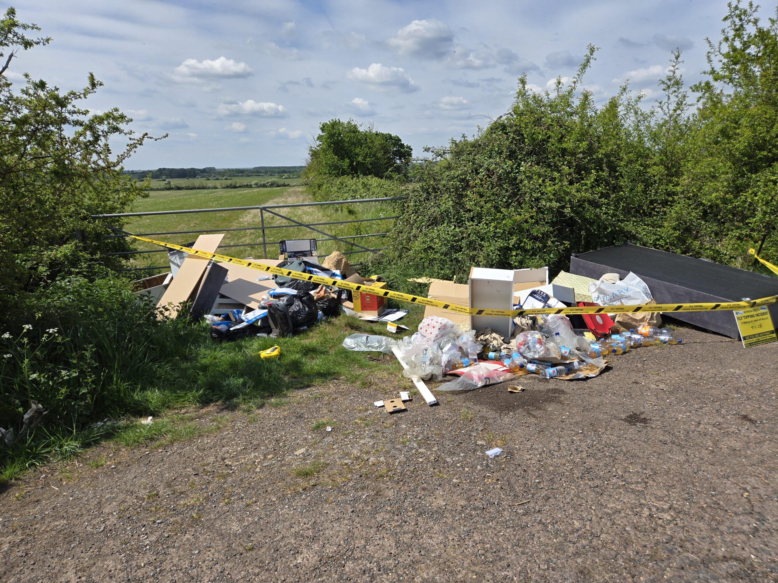 A wide shot of a fly-tipping incident on a rural gravel turnout next to a metal gate and green fields. A large pile of rubbish, including broken white furniture, cardboard boxes, plastic bags, and dozens of plastic bottles containing amber liquid, is cordoned off with yellow 