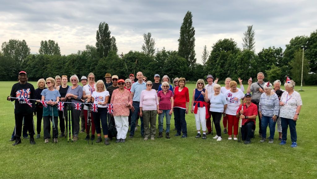 A group of people standing on a playing field. There are tall trees behind them. They are smiling for the camera and some are holding jubilee bunting