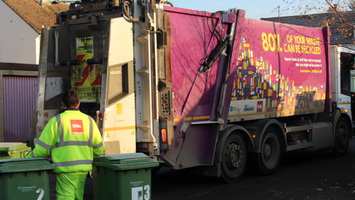 A photo of a bin man pulling two green wheelie bins to a waste and recycling truck