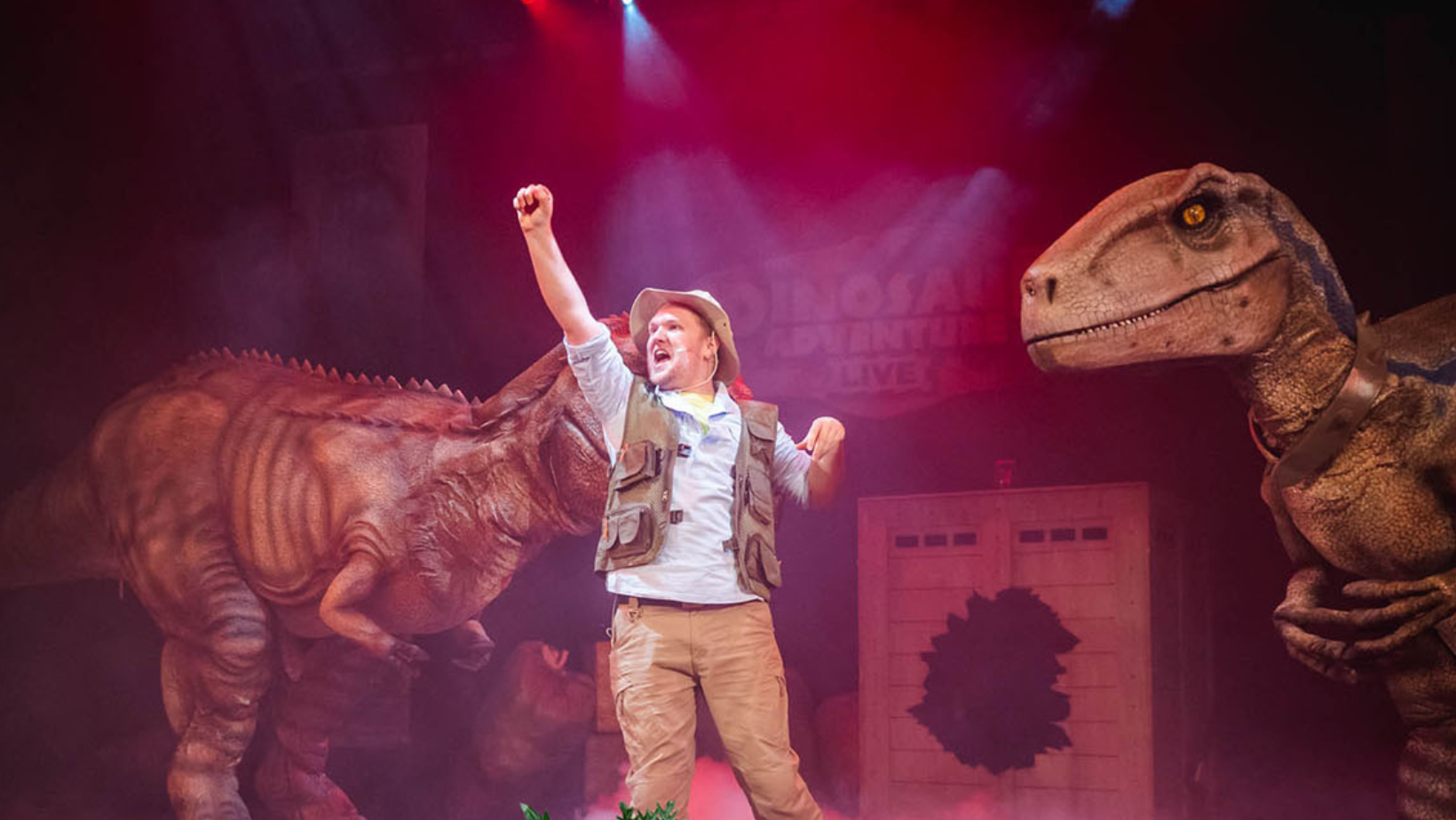 A picture may in explorers clothes with his fist in the air standing by two dinosaurs